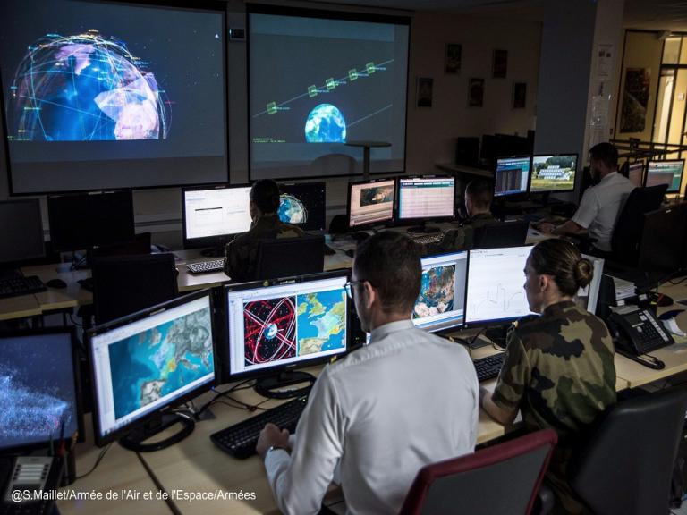 deux personnes sont devant leurs ordinateurs au COSMOS, dans la salle opérations. On peut distinguer des vues aériennes de la Terre et des trajectoires de satellites